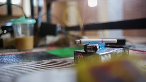 primer plano de cigarrillos en paquetes de cigarrillos y encendedores de gas con hermosas colas de humo con un fondo de un vaso de café con leche y cables esparcidos en la habitación de los trabajadores