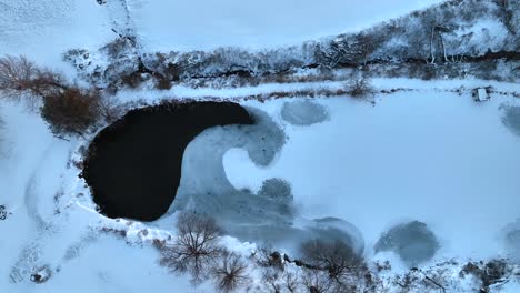 Von-Oben-Nach-Unten-Luftaufnahme-Eines-Bauernhofteichs,-Der-Teilweise-Zugefroren-Und-Mit-Winterschnee-Und-Eis-Bedeckt-Ist