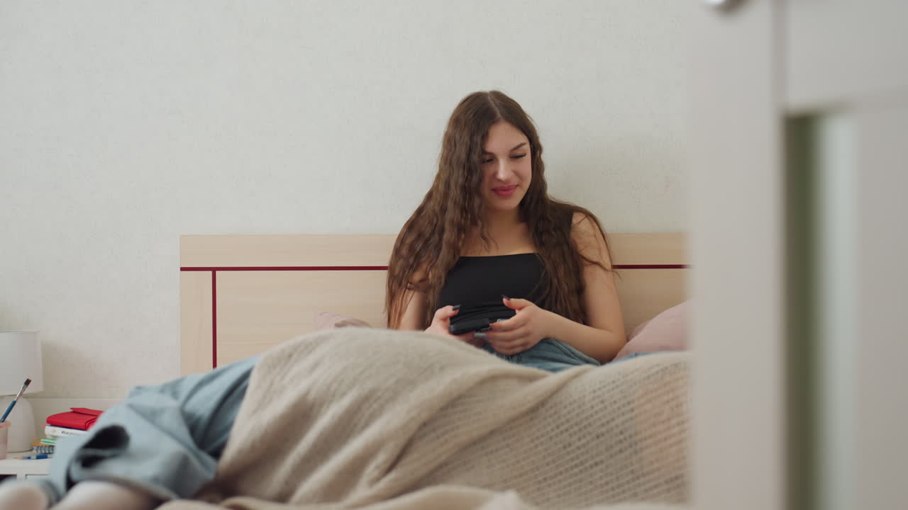 Young Lady Starts Day With Bright Smile, Female Sitting Comfortably In Bed Using Her Phone Peacefully, Cheerful Young Woman Begins Her Day By Relaxing In Bed With Warm Beverage And Her Phone