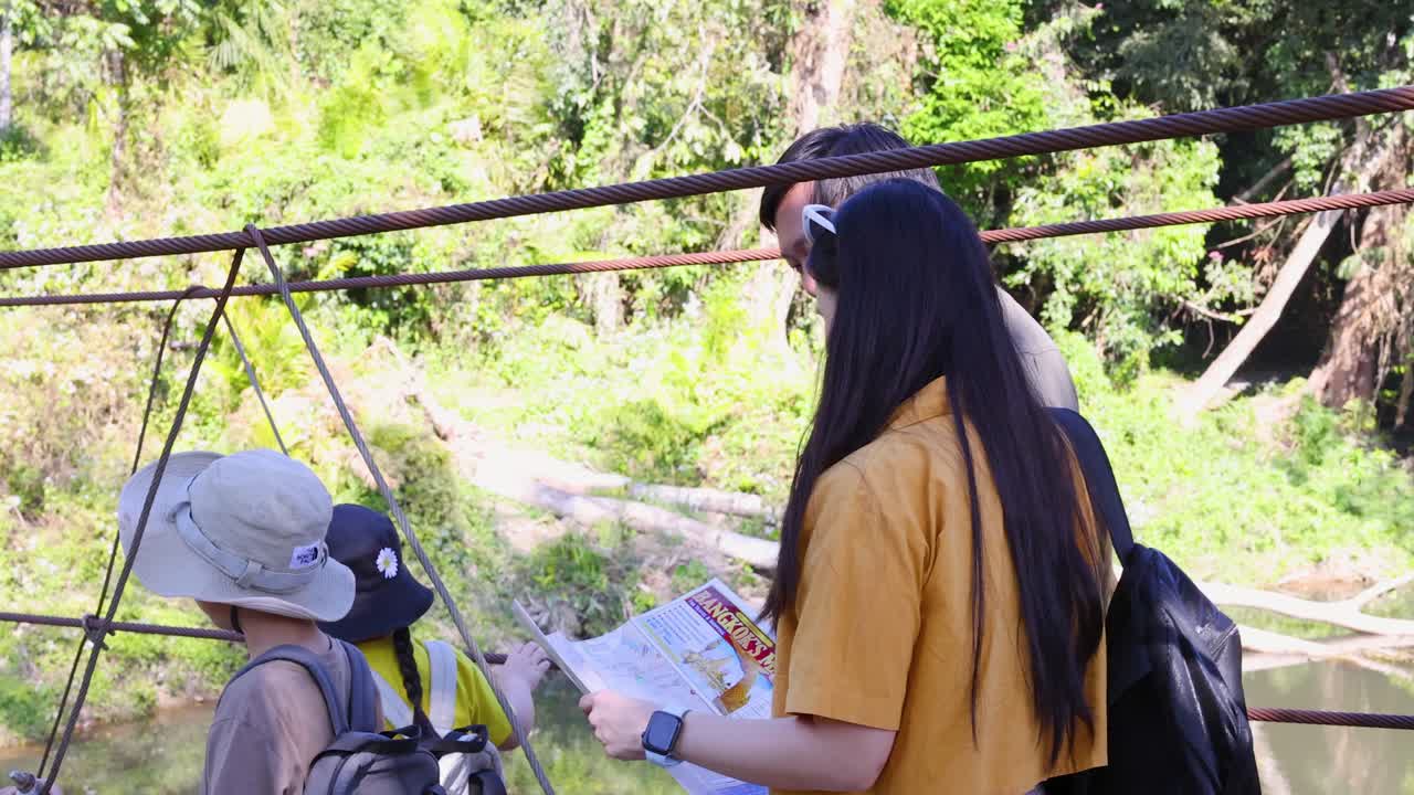 Família tailandesa feliz com crianças examina um mapa enquanto está em uma ponte suspensa na selva