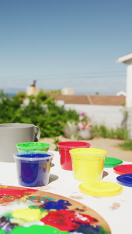 vertical video of close up of colourful paints and painting equipment lying on table in garden