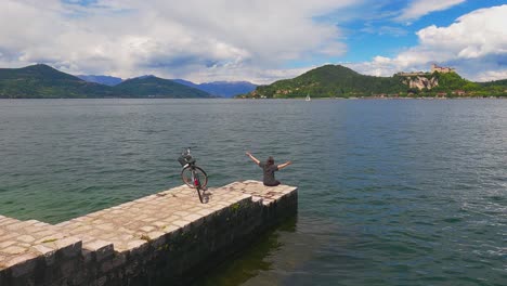vista posterior de una mujer feliz y despreocupada con los brazos abiertos y en bicicleta en el borde del embarcadero del lago maggiore, italia