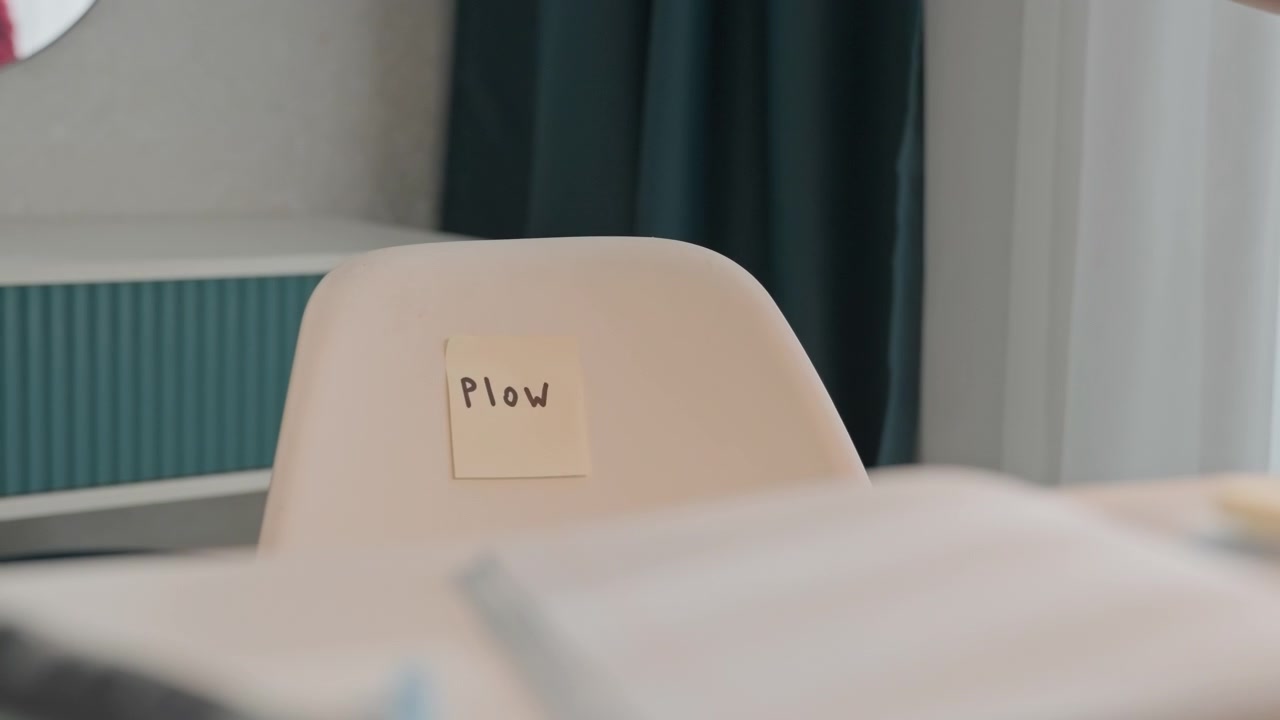 Mujer con blusa roja coloca notas adhesivas etiquetadas como 'Pieza'. La mujer alisa con ambas manos el respaldo del asiento, asegurándose de que quede bien. La escena transcurre en interiores con paneles de color verde azulado, cortinas oscuras y luz.