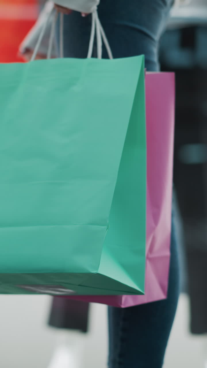 primer plano de bolsas de compras verdes y rosas sostenidas por una mujer en vaqueros y una camiseta blanca de manga larga, de pie frente a una moderna exhibición minorista con maniquíes y un fondo rojo vibrante