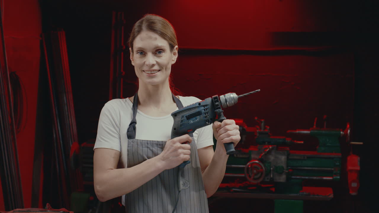 앞치마를 입은 젊은 여성의 초상화는 금속 작업장에서 전기 드릴을 켜고 카메라를 보고 웃고 있습니다.