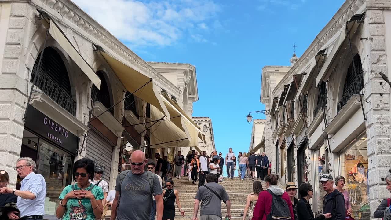 People walking up the steps of Rialto Bridge on a sunny day in Venice lined with stores