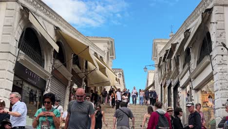 people walking up the steps of rialto bridge on a sunny day in venice lined with stores