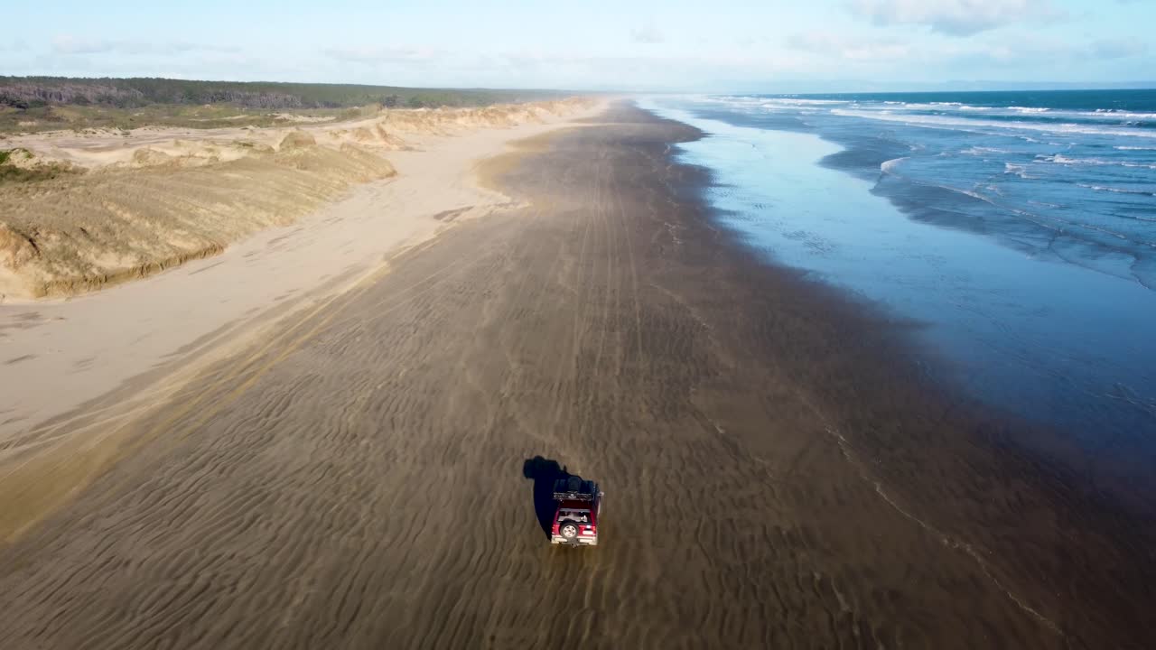 Drone view of offroad 4 wheel drive red car with rooftop tent driving on sand next to the ocean on a long beach on a sunny day at 90 Miles Beach, Northland, New Zealand.