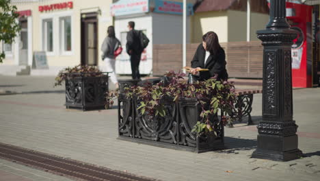 white girl seated in public space flipping through pages of book thoughtfully, with background showing flower pot swaying in the wind, pedestrians walking, and vending machine nearby