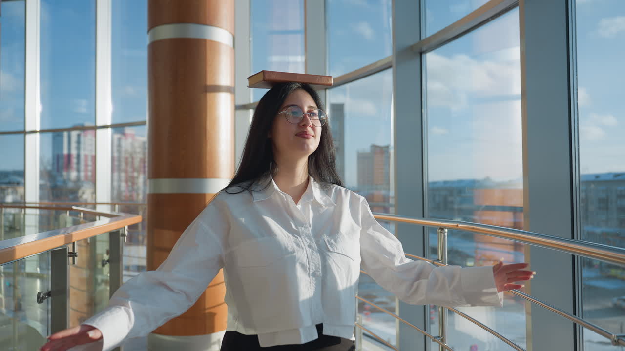Young student playfully walking with arms stretched and book balanced on head in sunlit modern hallway, wearing glasses and white shirt, surrounded by large glass windows