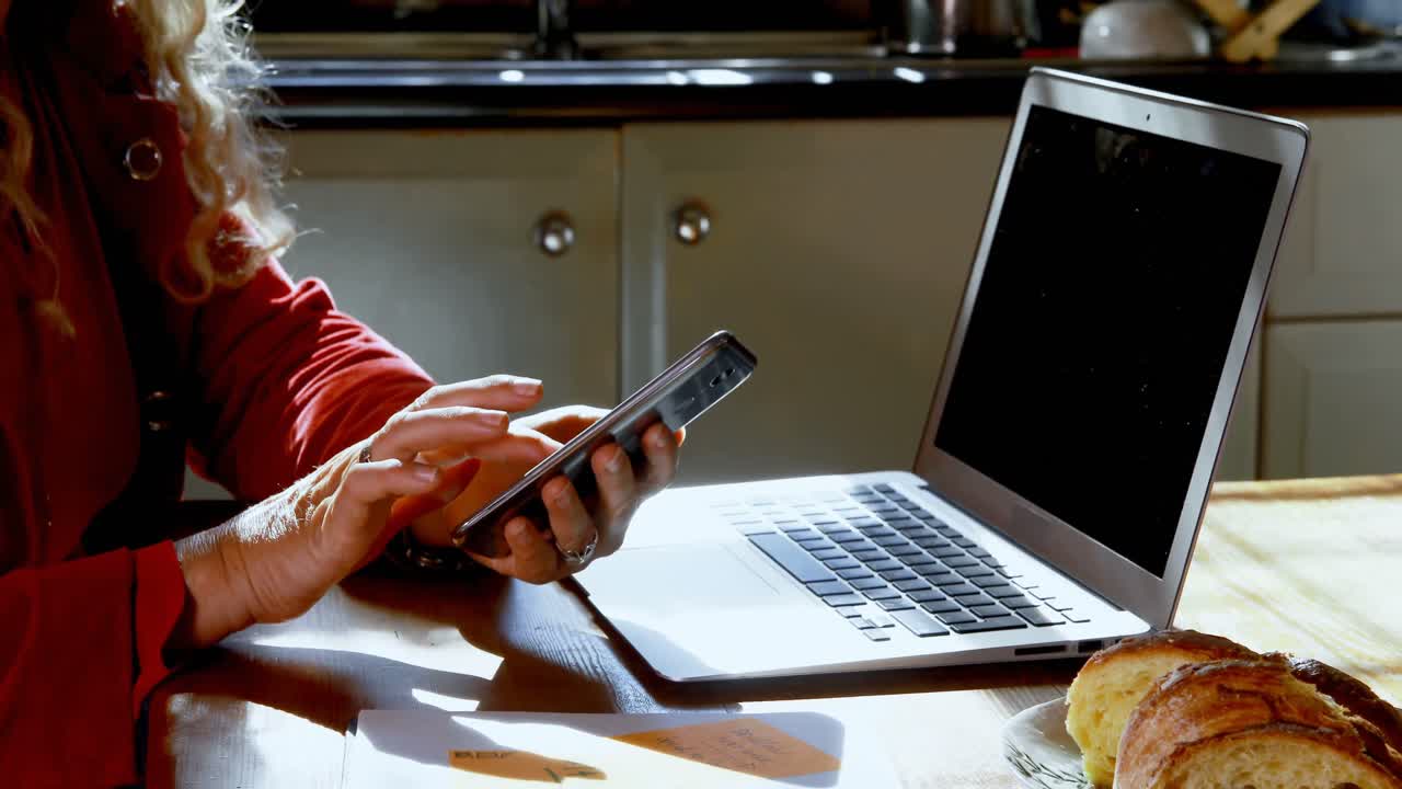 mujer madura usando teléfono móvil y portátil en la cocina en casa 4k