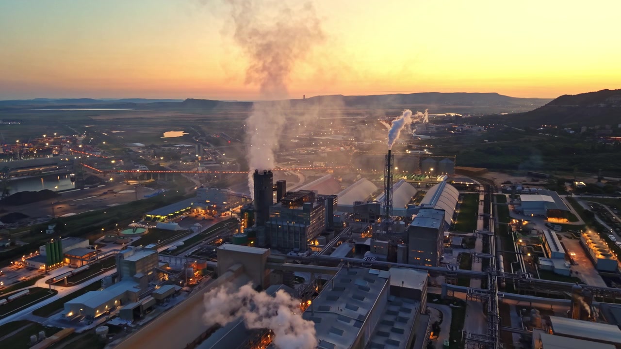 Vista serale di una grande area industriale con fumo che sale da edifici e macchinari al tramonto