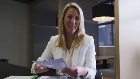 Caucasian-businesswoman-having-video-chat-going-through-paperwork-in-modern-office