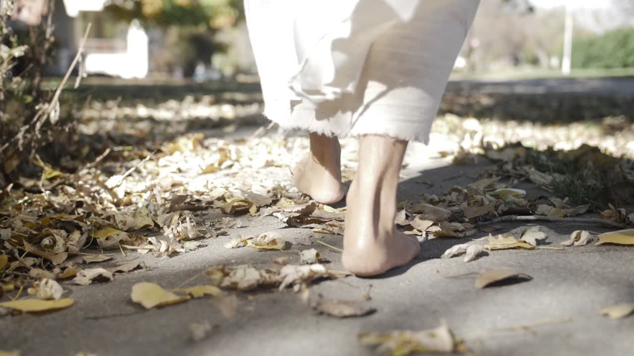 A dramatic slow motion closeup of Jesus' feet walking down a sidewalk while he wears a white, tattered robe with the light of the sun shining on him