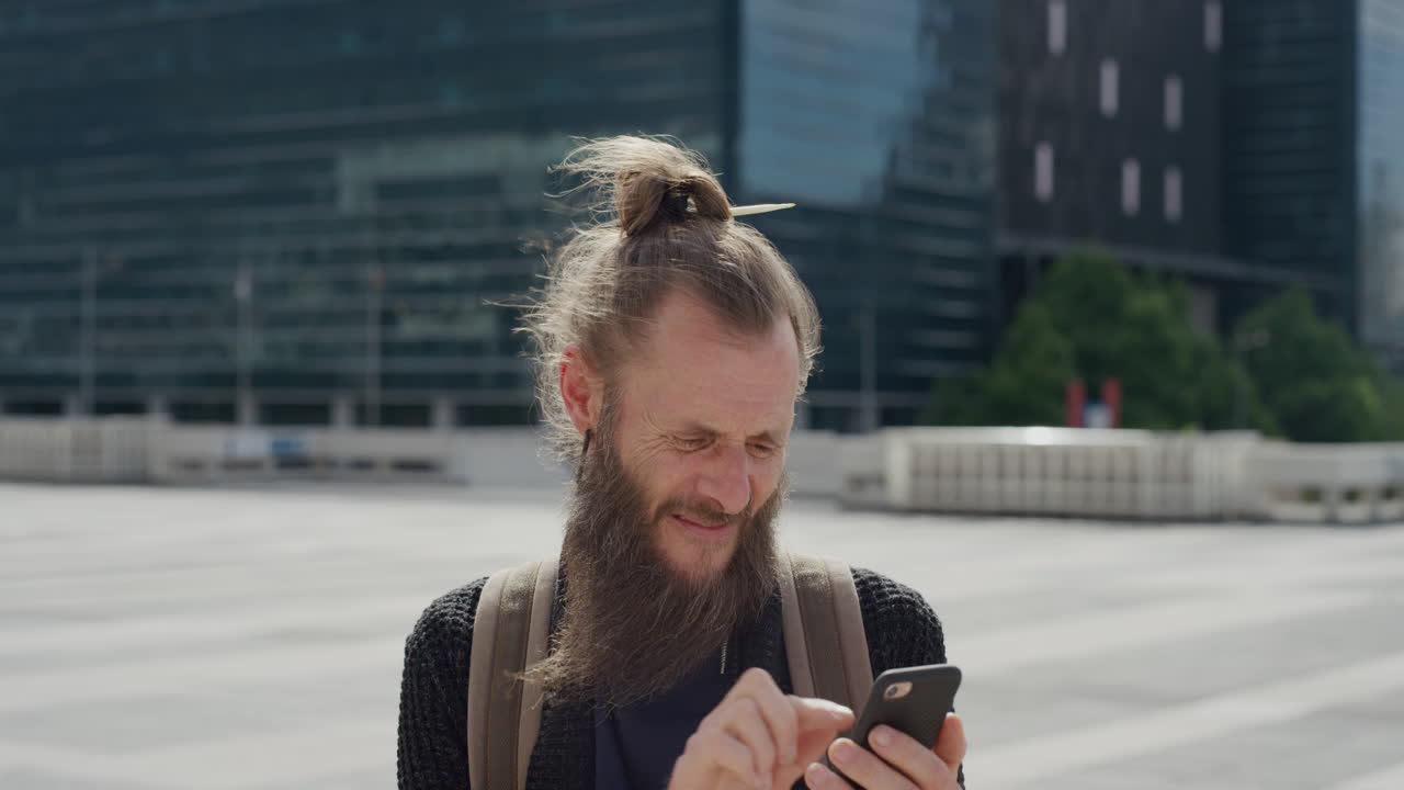 portrait mature bearded hippie man using smartphone in city enjoying texting browsing online on warm sunny urban day wind blowing