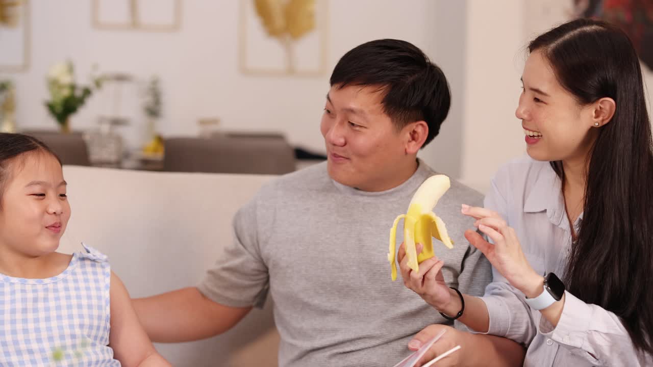 A joyful family shares a moment eating bananas in a cozy living room setting with warm lighting