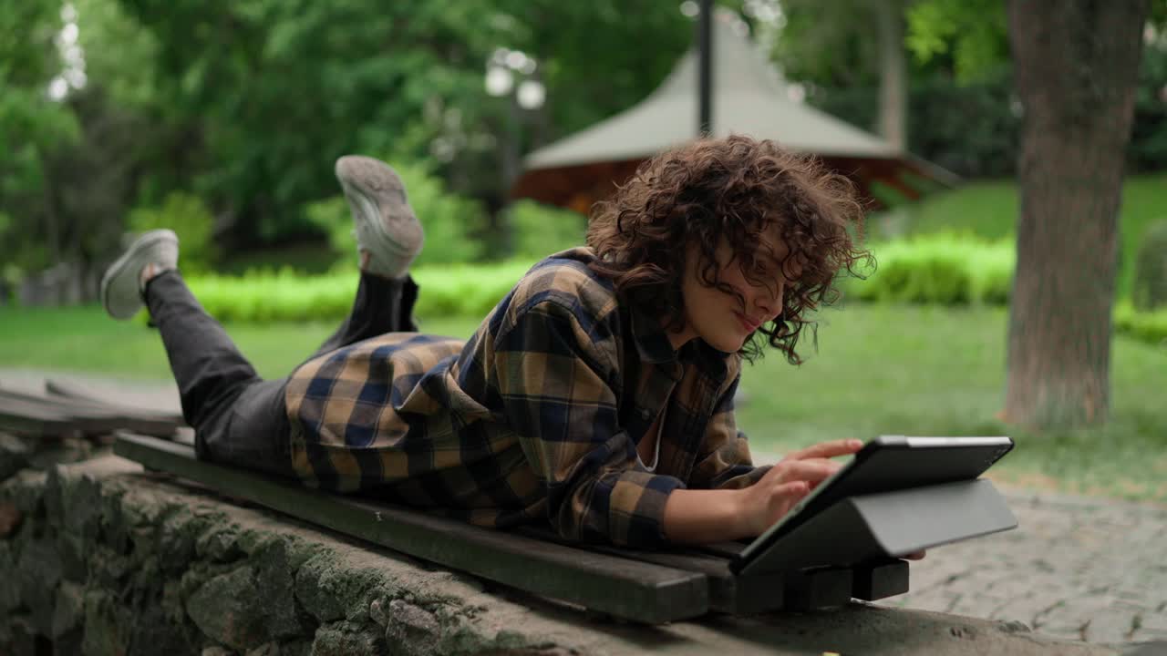 Happy girl student with curly hair in a checkered shirt uses a tablet and lies on a bench in the park