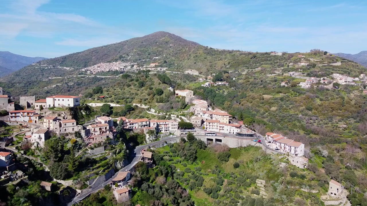Ripresa aerea in allontanamento del borgo medievale collinare di Savoca in Sicilia, circondato da lussureggianti montagne mediterranee, con il Bar Vitelli in Piazza Fossia, famoso come location delle riprese de Il Padrino