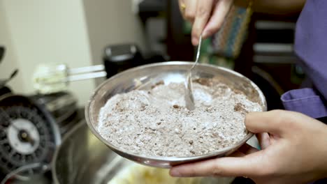 person mixing flour and cocoa powder in a metal bowl with a spoon. slow motion