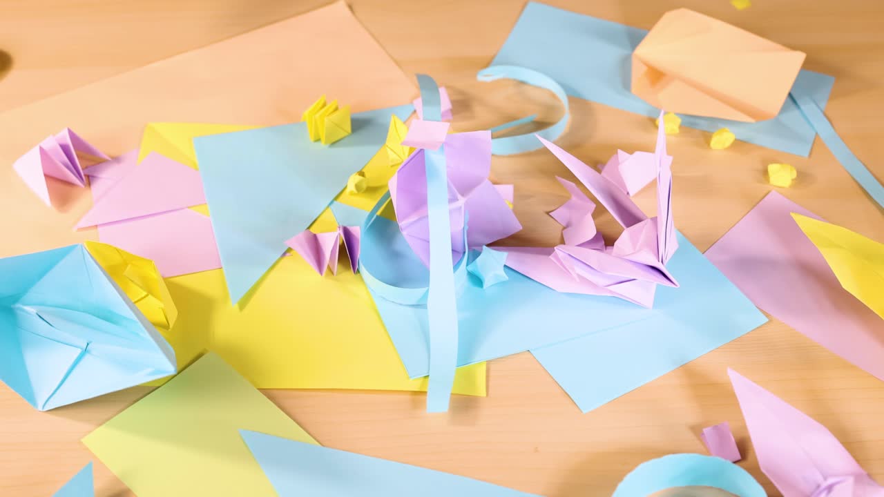 Overhead view of hand folding pastel origami birds and boats on wooden table, bright lighting