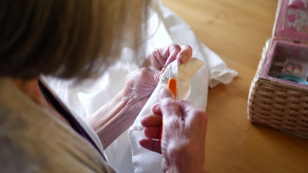 una anciana cortando el hilo de un viejo botón con unas tijeras en una camisa de vestir blanca mientras repara la prenda con su kit de costura