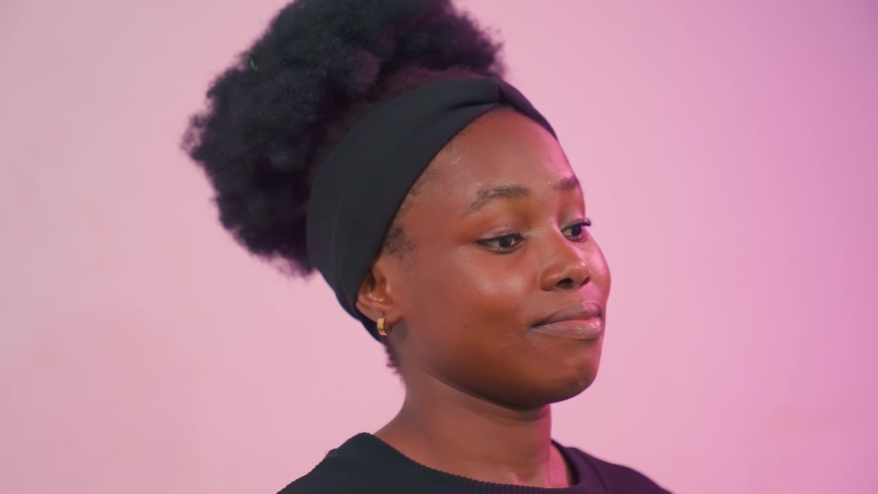 Primo piano di un ritratto in studio di profilo pensieroso di una madre nera. Giovane donna nera con afro e fascia per capelli su sfondo rosa, espressioni sottili che passano da pensierose a serene mentre si gira di profilo.