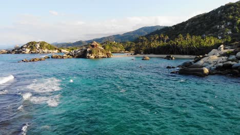 Aerial-view-low-over-waves-and-shallow,-blue-sea,-on-a-rocky-coastline,-in-Tayrona-national-natural-park,-during-golden-hour,-in-the-Caribbean-region-of-Colombia---dolly,-drone-shot