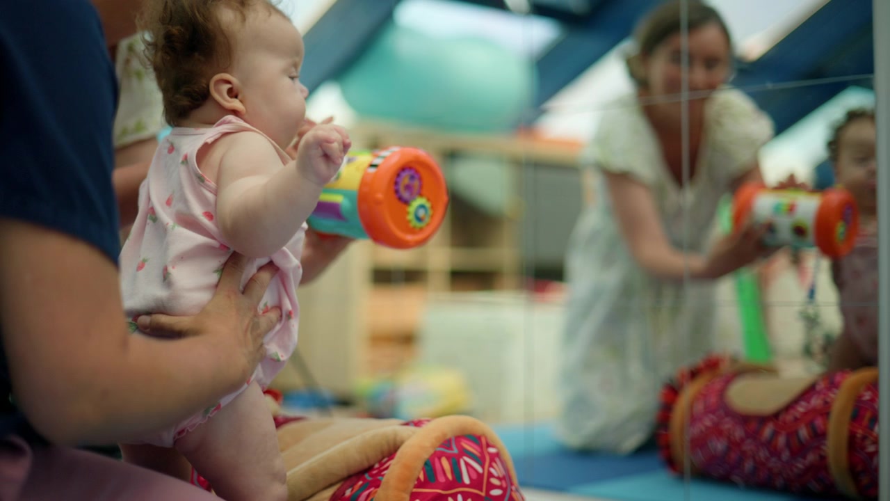 Bebê praticando ficar em pé e segurando um brinquedo colorido durante uma sessão de fisioterapia, auxiliado por uma pessoa que oferece apoio.