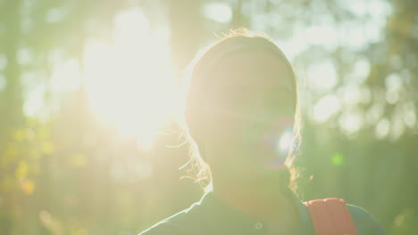 lady in green shirt and hair packed behind stands in forest adjusting her bag with warm, gentle smile, sunlight filters through trees, illuminating her hair with radiant glow