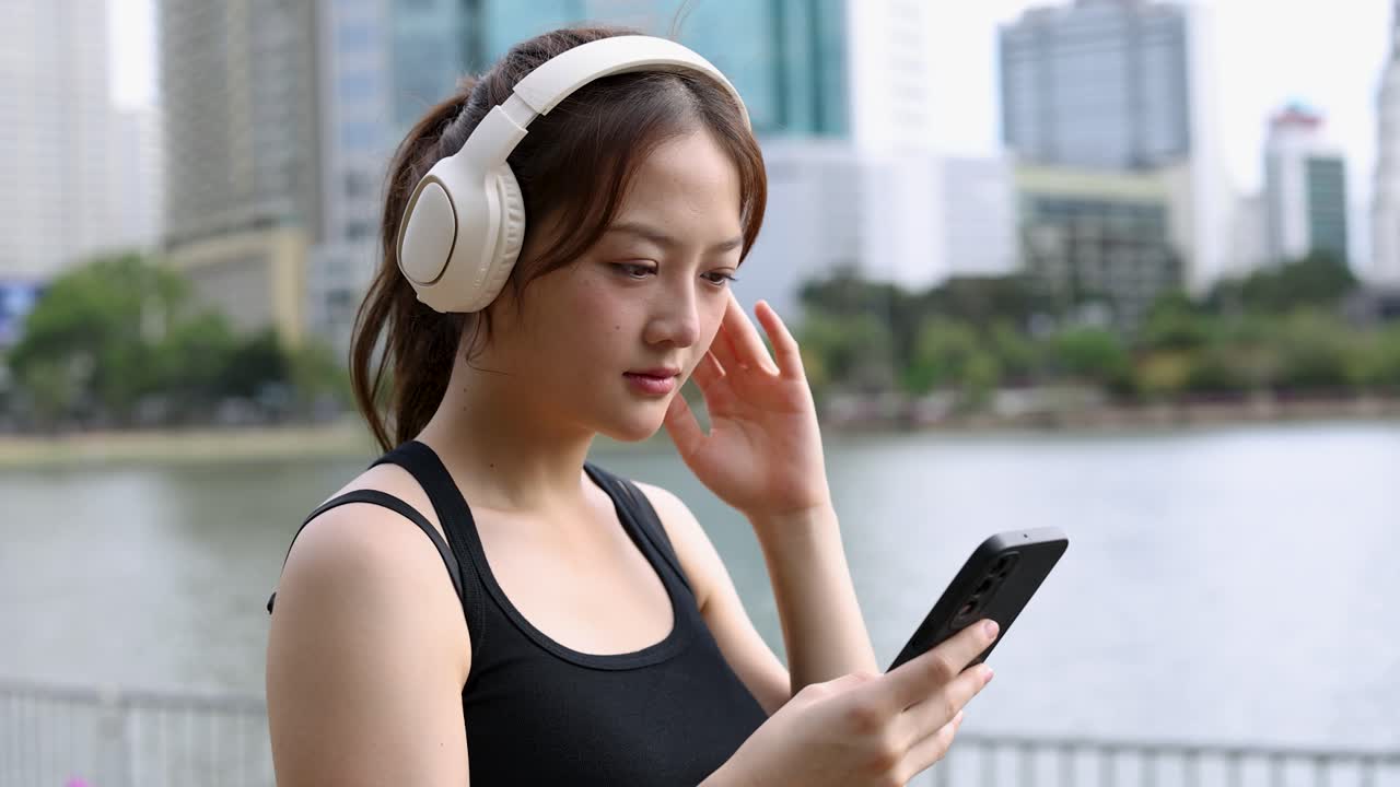 Donna asiatica in abbigliamento sportivo usa uno smartphone e cuffie accanto a un lago cittadino