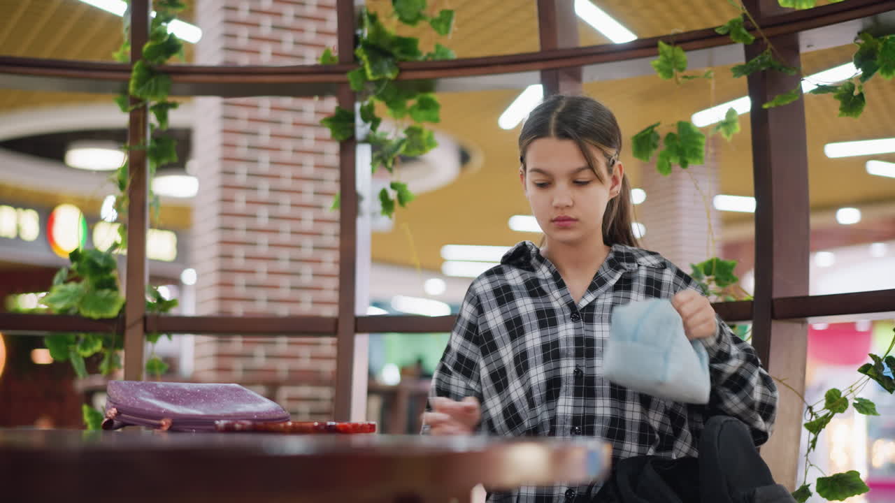 joven con una expresión preocupada saca un espejo y artículos personales de su bolsa, examinando su cabello mientras está sentada en un espacio interior acogedor con plantas verdes y iluminación cálida a su alrededor