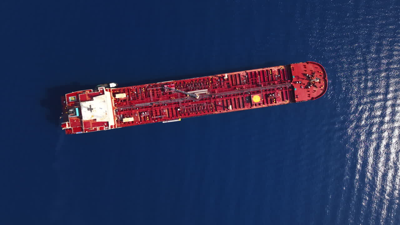 Drone executes a top-down orbit around a stationary red oil tanker in deep blue water near a Sicilian oil refinery