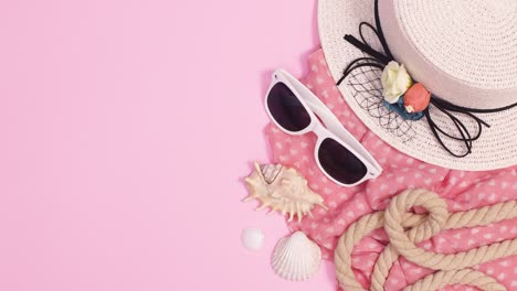 summer beach hat, sunglasses and scarf appear with shells and rope on pastel pink background. stop motion