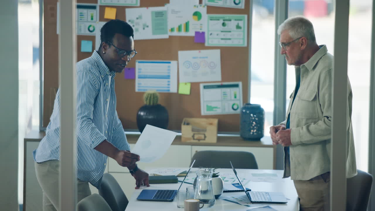 Two Businessmen Discussing Documents and Laptops in a Modern Office