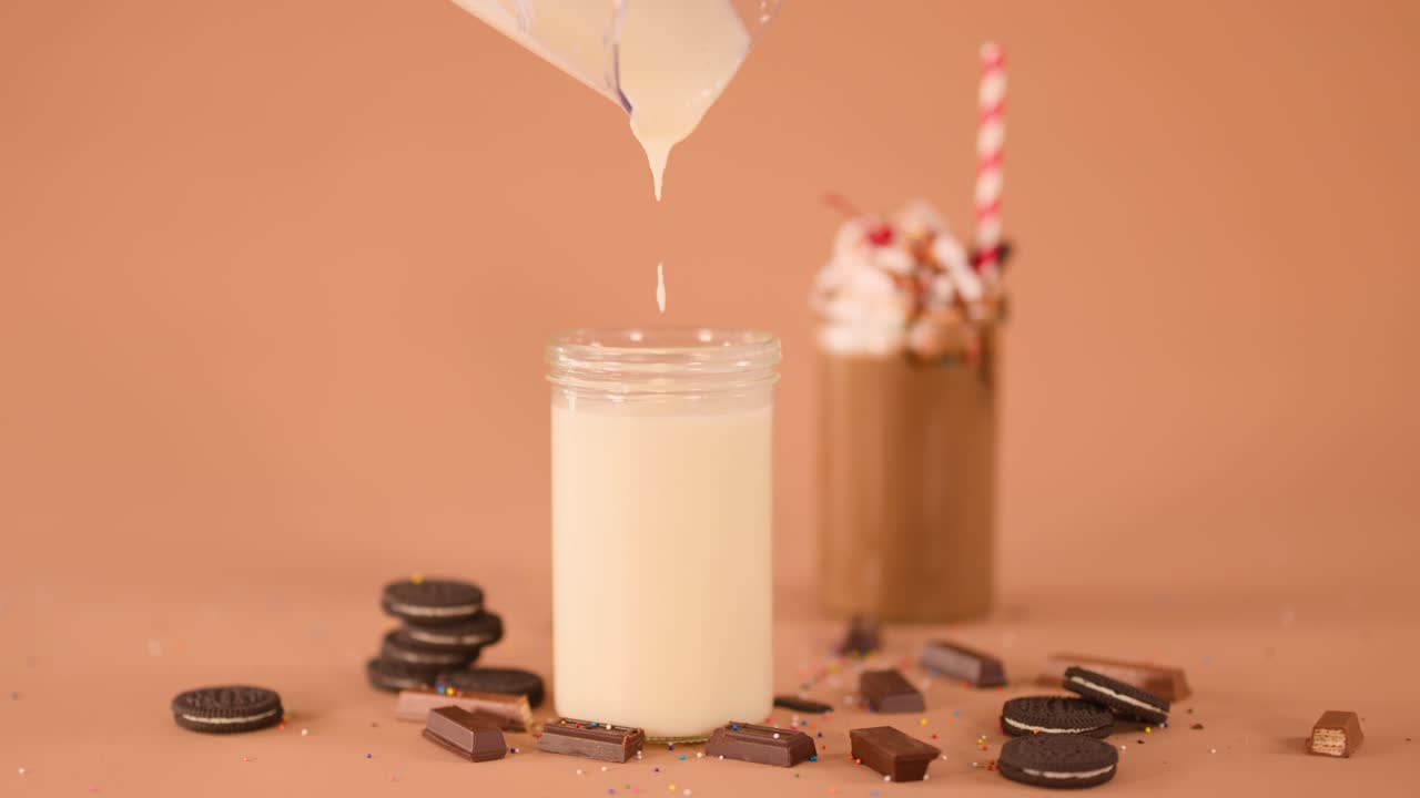 Thick creamy liquid pours into glass beside decorated chocolate milkshake, cookies, and candy pieces