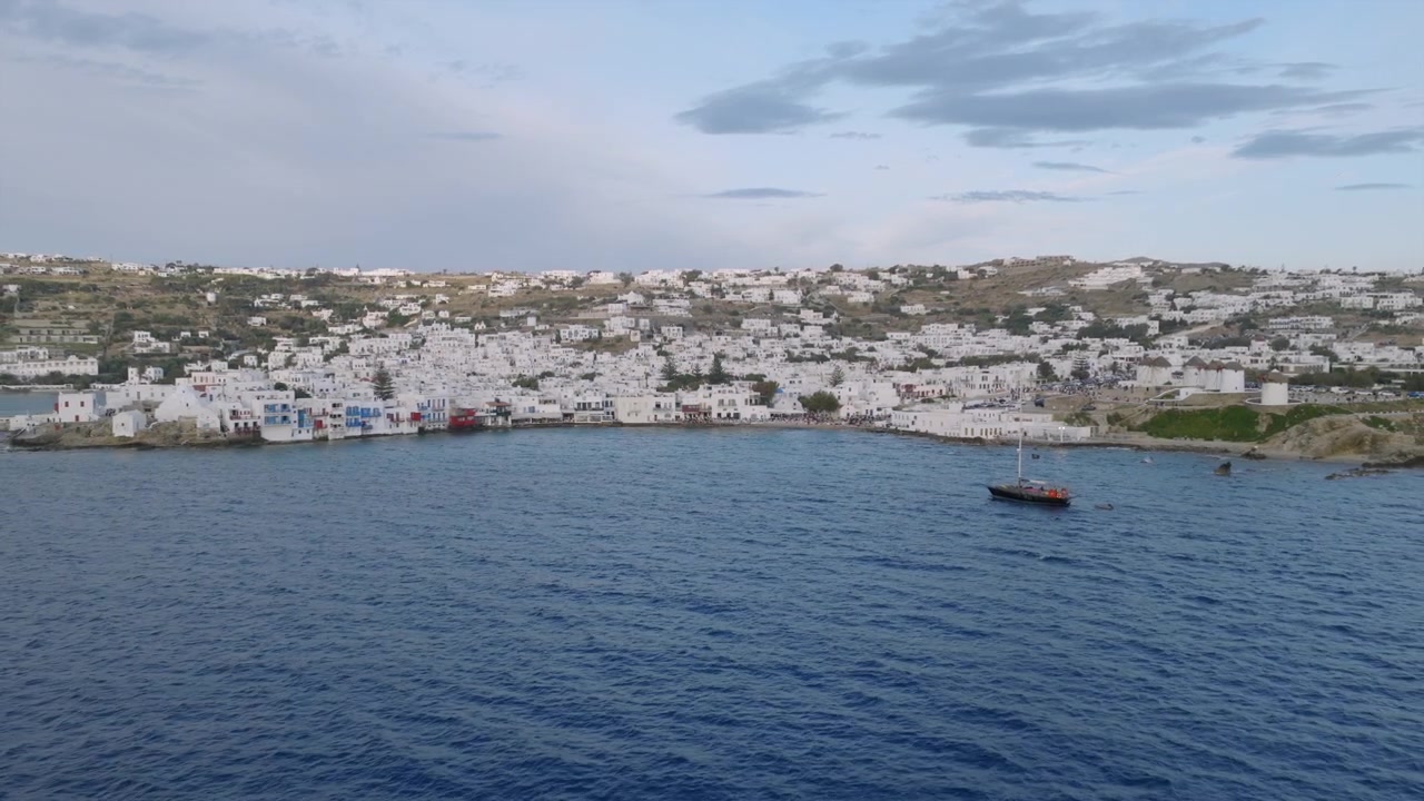 Ripresa aerea con drone che si avvicina alla città di Mykonos attraverso acque di un blu intenso, con una barca a vela ancorata al largo ed edifici bianchi che coprono le colline costiere