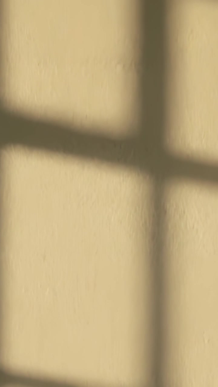 Vertical video: Sunlight shifting, soft multi-pane window shadow drifting across beige wall at home