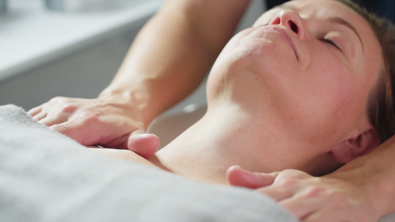 Skin therapist rubbing head and neck of model lying relaxed on massage chair in spa with white background during tranquil facial therapy session showcasing skilled hands and serene environment