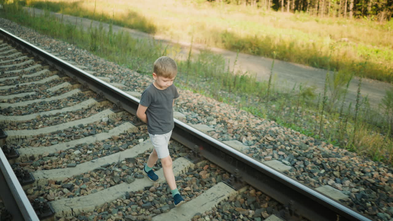 side view of thoughtful boy walking quietly on railway track with hands behind his back, dressed in casual outfit, surrounded by rural scenery with gravel stones and dry grass in soft afternoon light