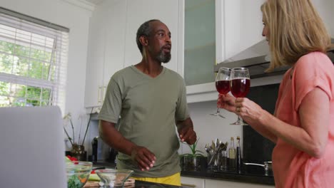 diverse senior couple preparing food in kitchen using laptop and drinking wine