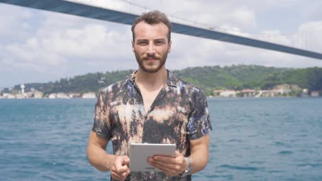 man using tablet by the sea laughs looking at camera.