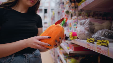 close-up of a hand picking a vibrant orange purse with yellow details from a store shelf, shopper examines stylish accessory among various packaged items, retail shopping for fashion or gift products