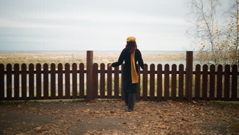 rear view of a lady in a yellow beret and black coat walking towards a wooden fence, resting her hand on it, admiring a serene river landscape with dry foliage scattered around