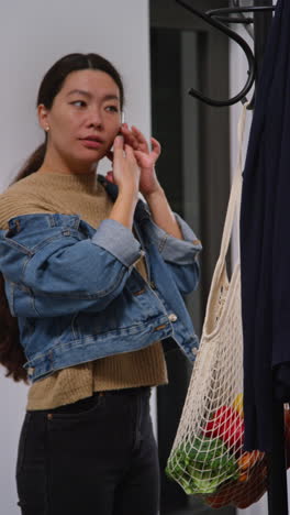 vertical video of woman returning from shopping talking on mobile phone hanging up coat on stand before carrying bag into home