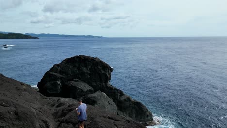 Cinematic-aerial-view-of-Male-Hiker-walking-through-basalt-boulders-revealing-stunning-vast-ocean-bay-in-Catanduanes,-Philippines