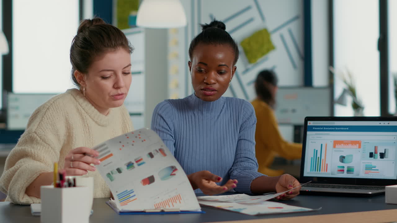 Team of colleagues analzying market share looking at laptop comparing sales charts on clipboard