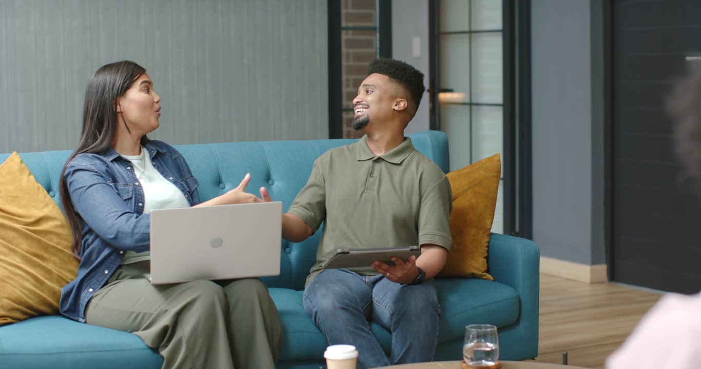 Diverse coworkers in lounge woman showing laptop after hair touch man with tablet nodding handshake