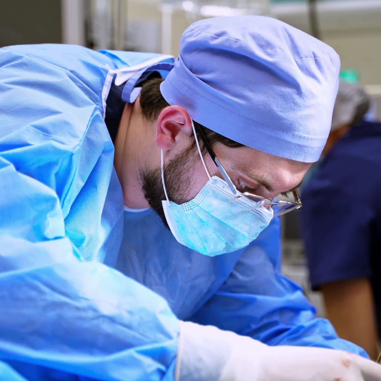 Surgeon in mask and glasses in hospital. Male specialist doctor in blue medical uniform operates with surgical tools in the operating room.