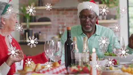 Animation-of-white-snowflakes-over-diverse-senior-friends-in-hats-saying-grace-at-christmas-dinner