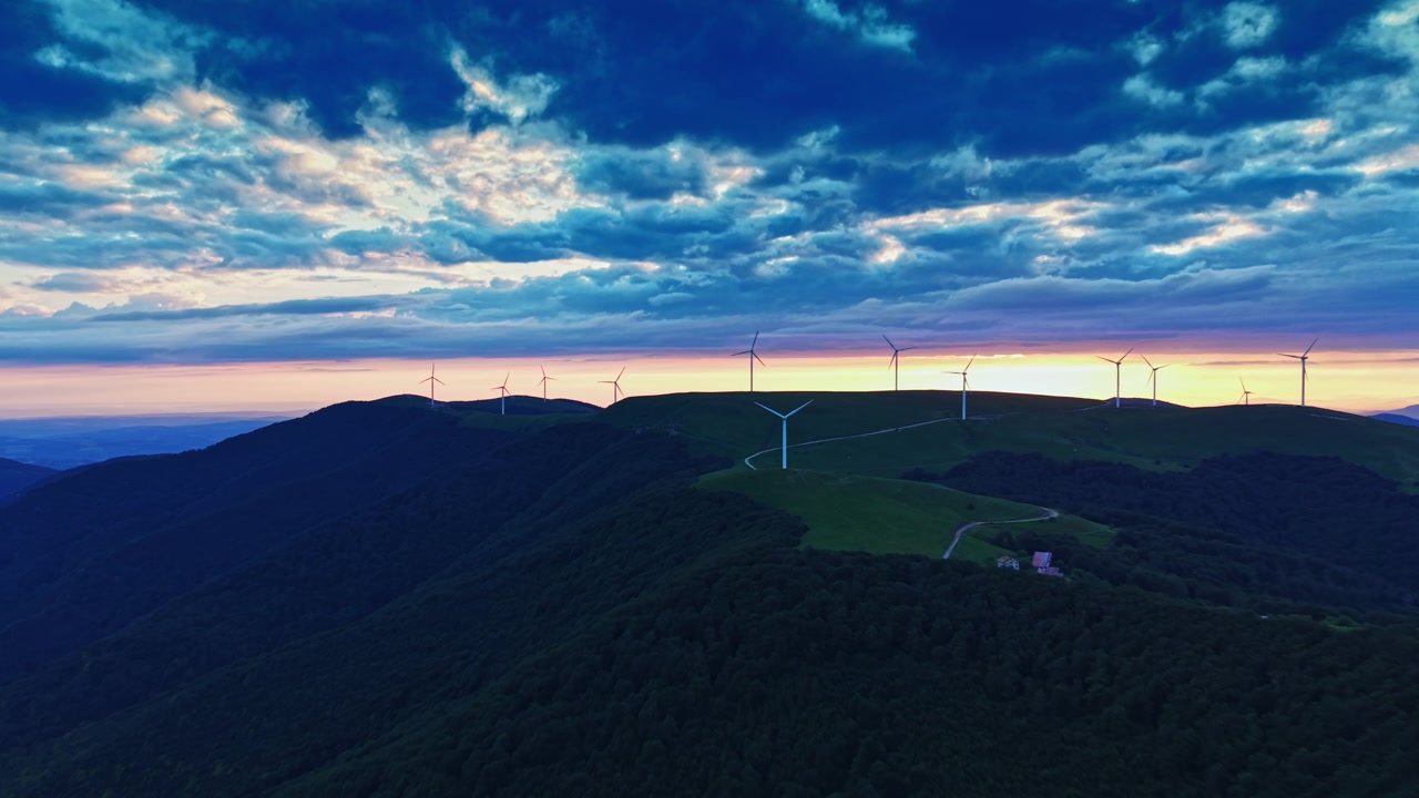 Tramonto su turbine eoliche in cima a una collina con nuvole drammatiche e terreno ondulato, visto dall'alto nel cielo serale.